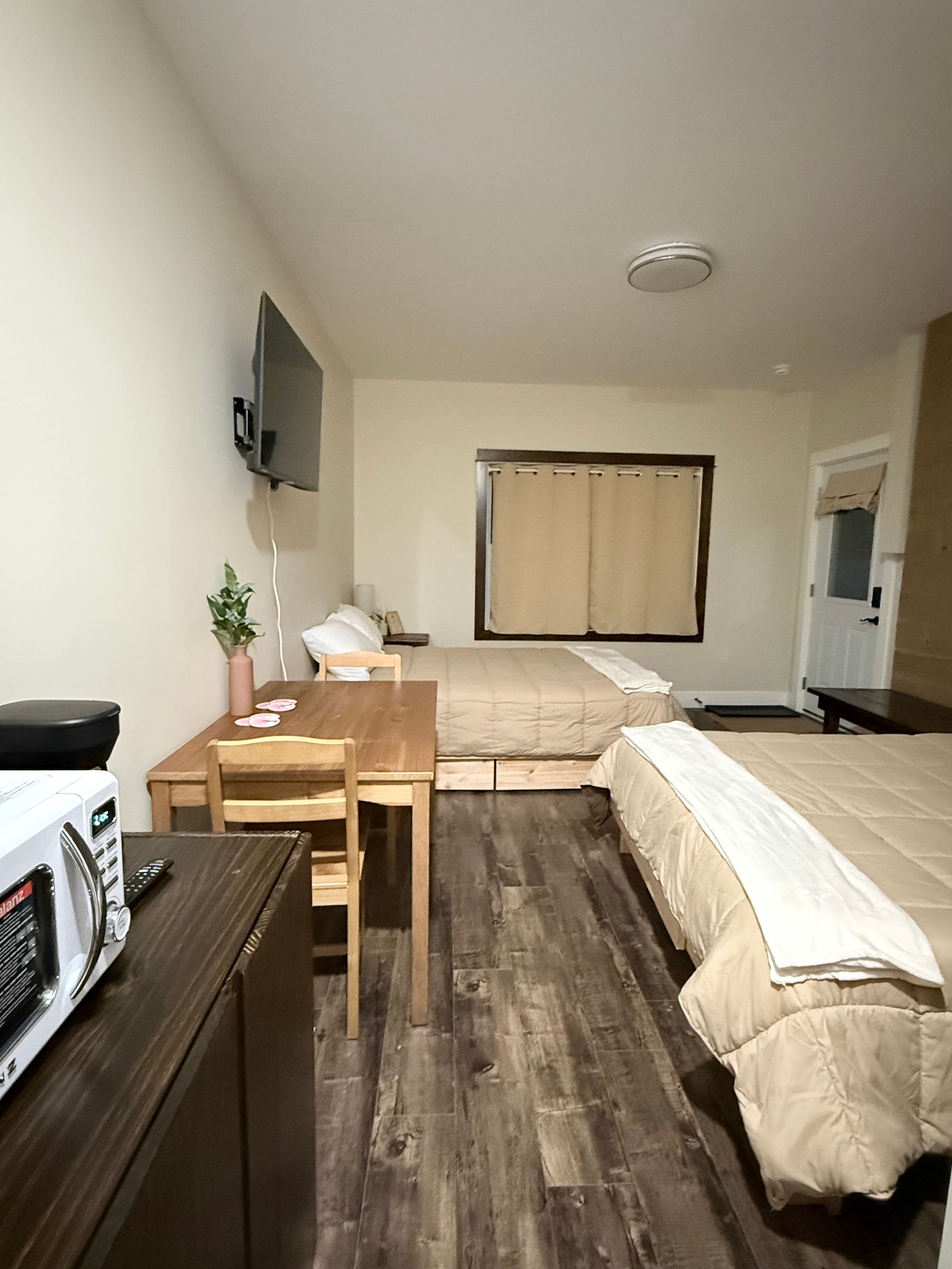 Small bedroom with two beds, a table, chairs, TV, and kitchenette area in a simple motel room.