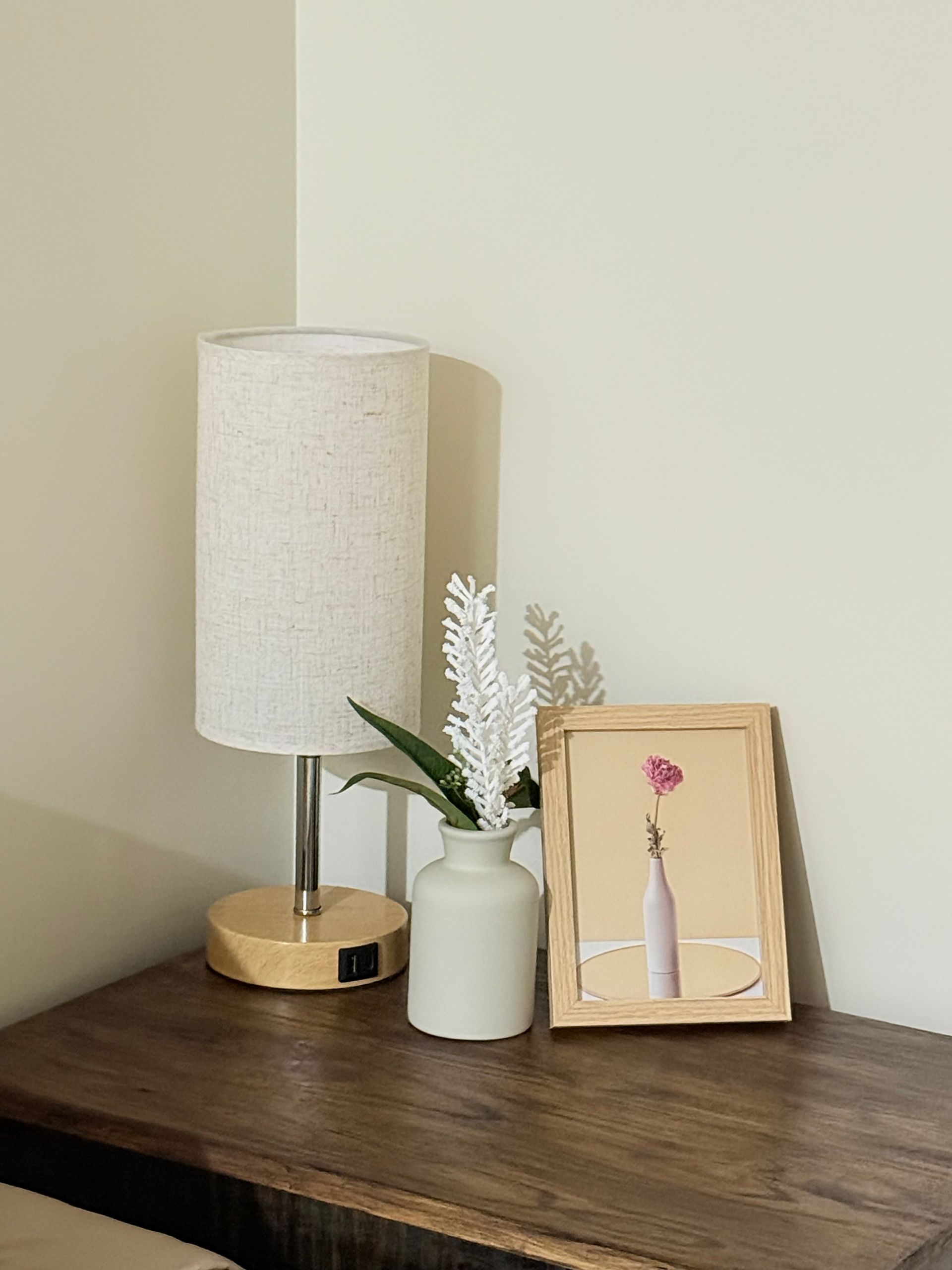 Small table lamp, white vase with dried flowers, and framed art on a dark wood side table.