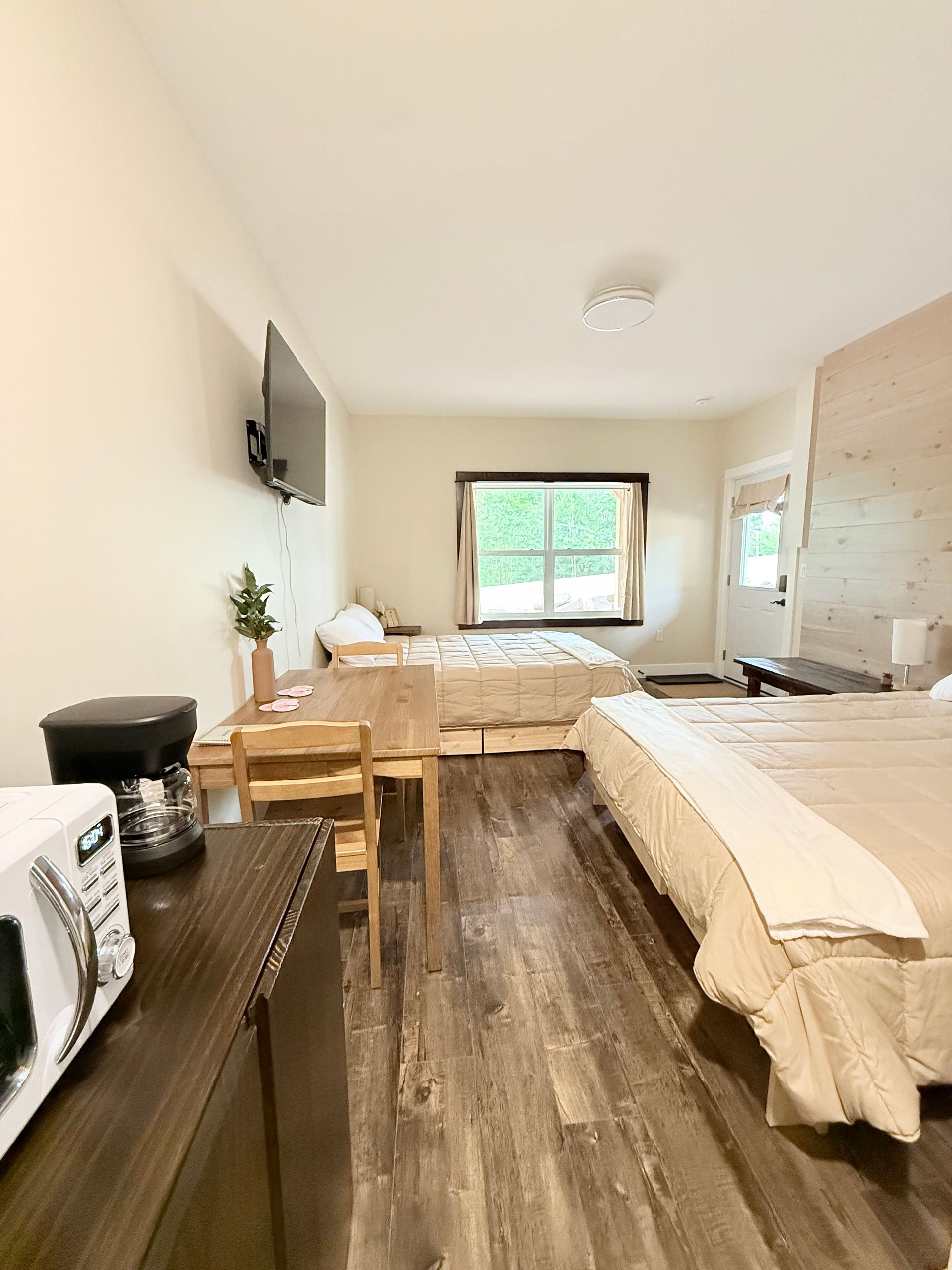 Small bedroom with two beds, a desk, wall TV, and window with greenery outside.