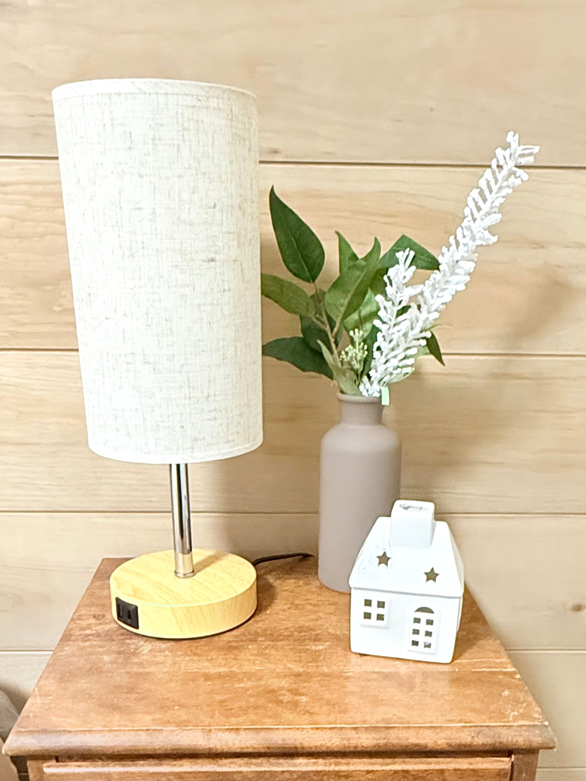 Small table lamp with white shade beside a vase of greenery and a small white house decor on a wooden surface