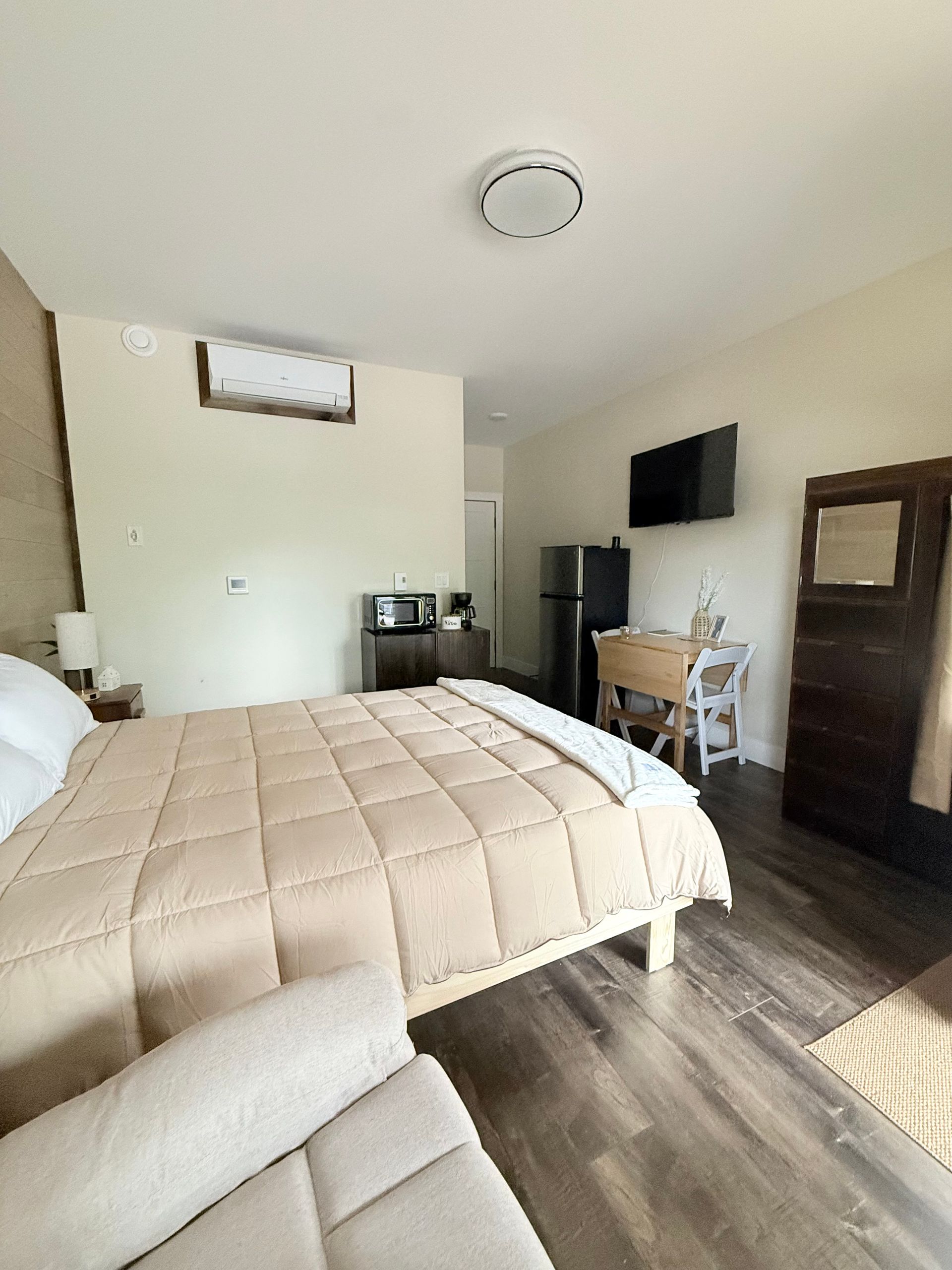 Small bedroom with a bed, wall-mounted TV, dresser, chair, and dark wood floor.