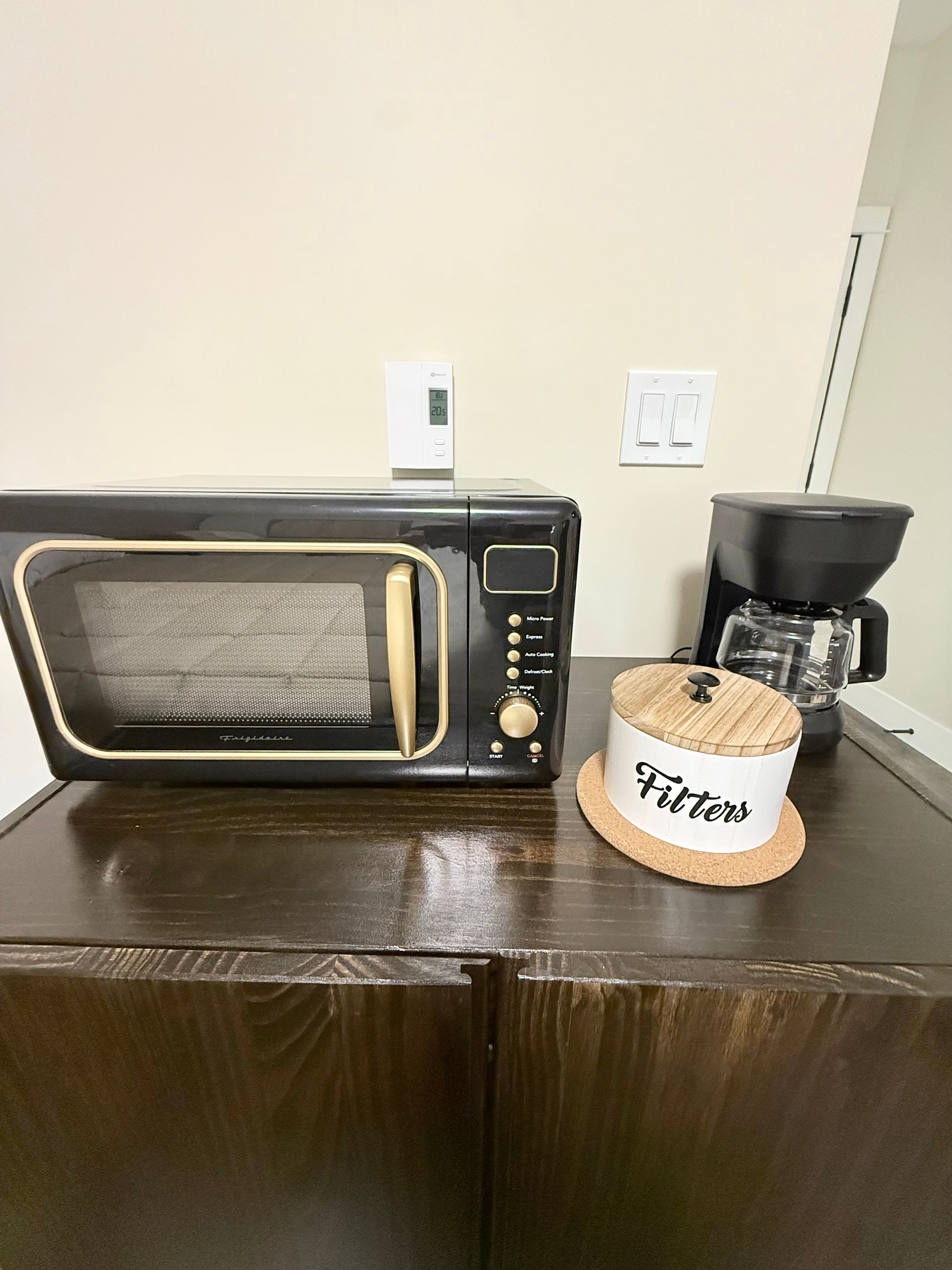 Countertop with a microwave, coffee maker, and a small jar of tea bags against a beige wall.