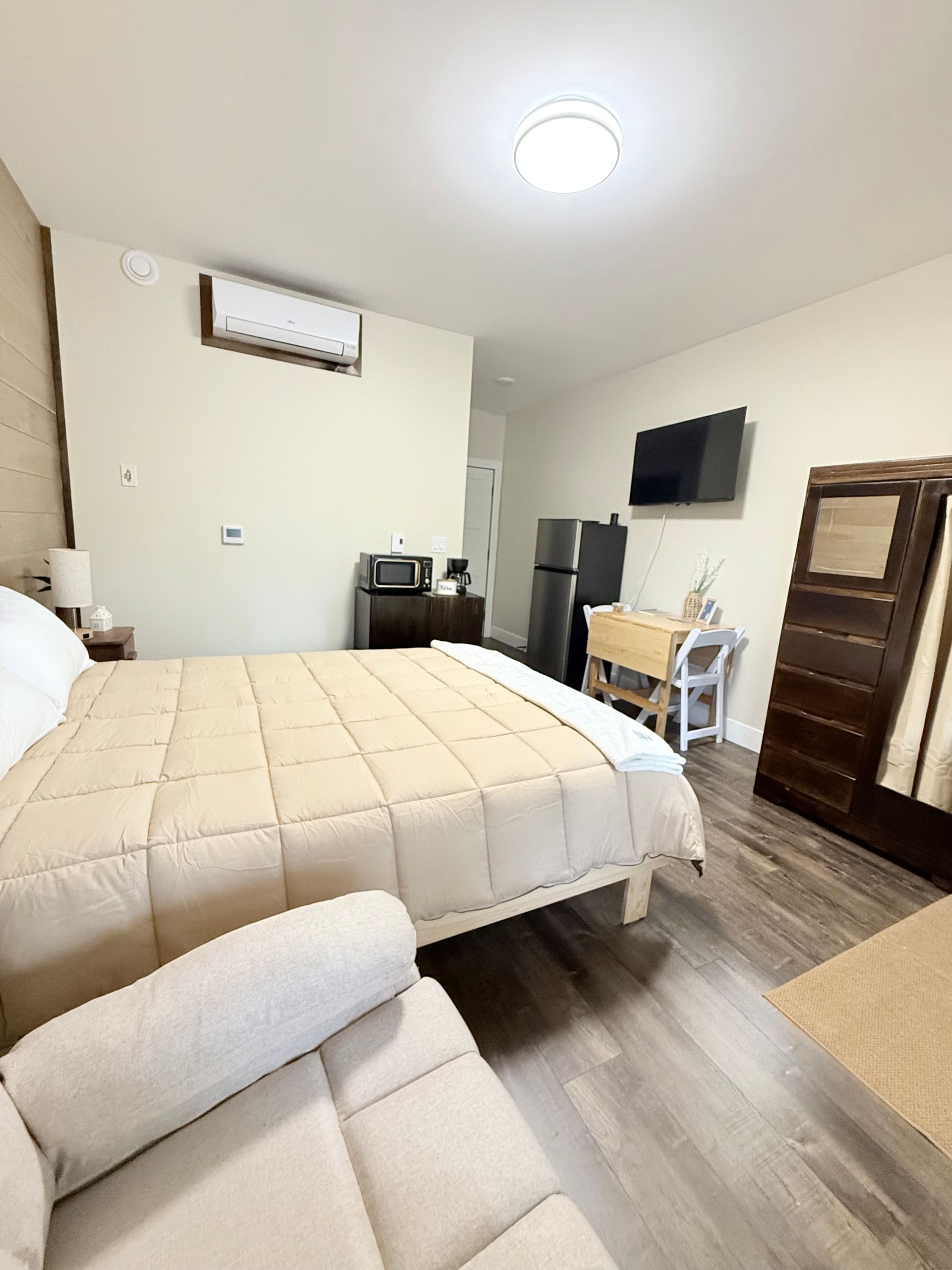 Bedroom with bed, wall-mounted TV, dresser, chair, and air conditioner in a bright room.
