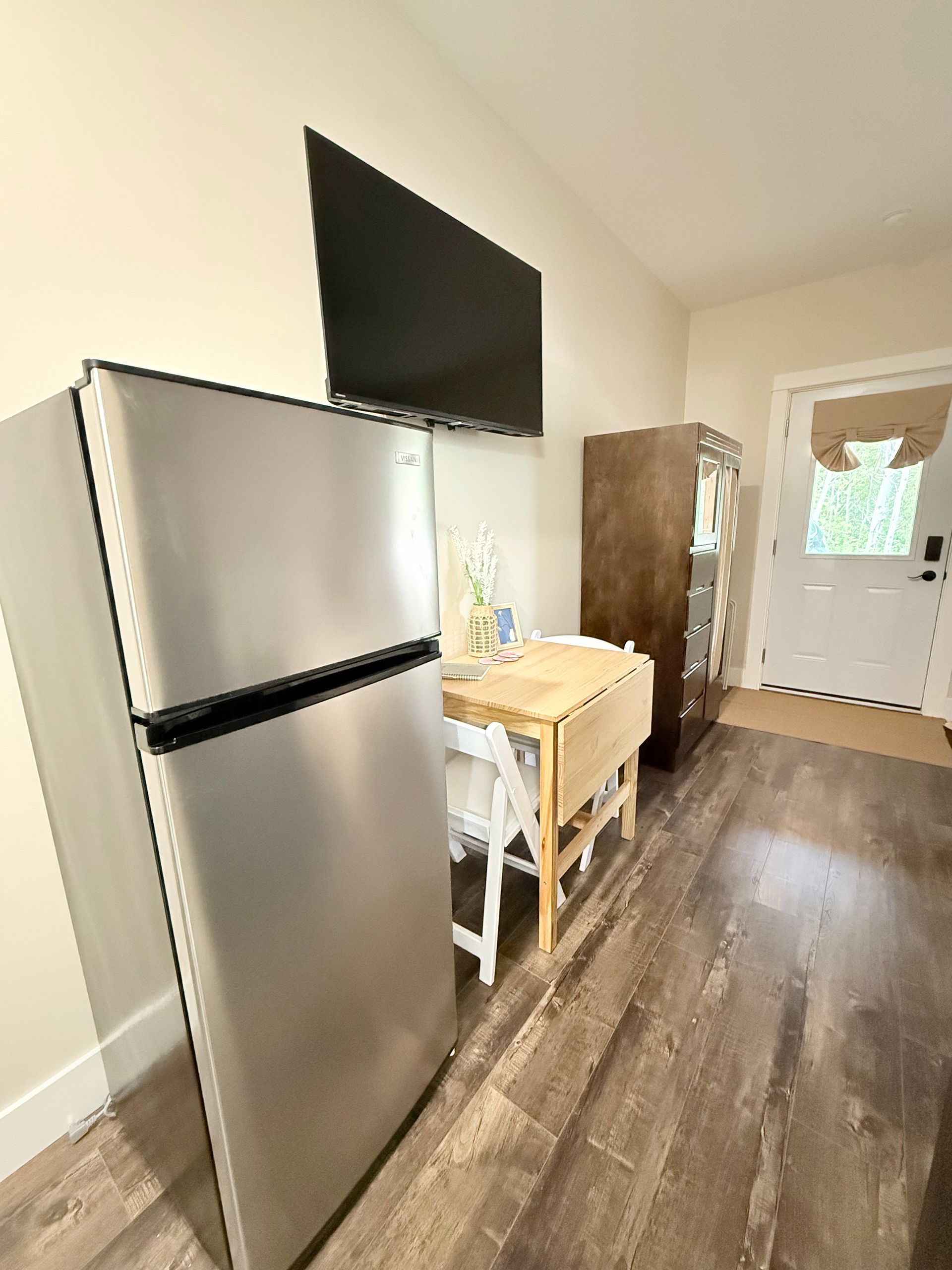 Small kitchen with stainless steel fridge, TV mounted above, and a compact table by the door.