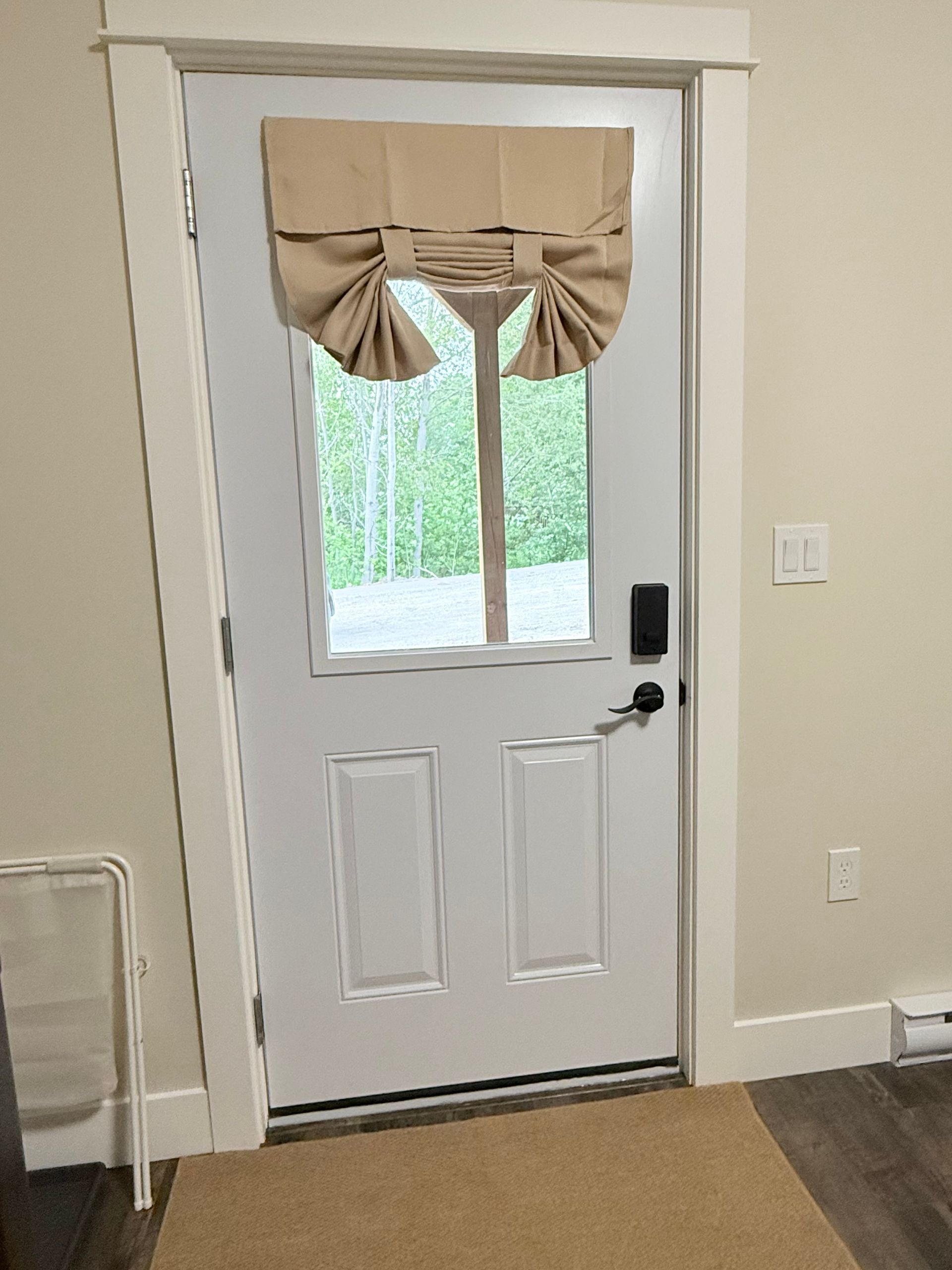 Front door with a small curtained window, white trim, and beige walls inside a home.