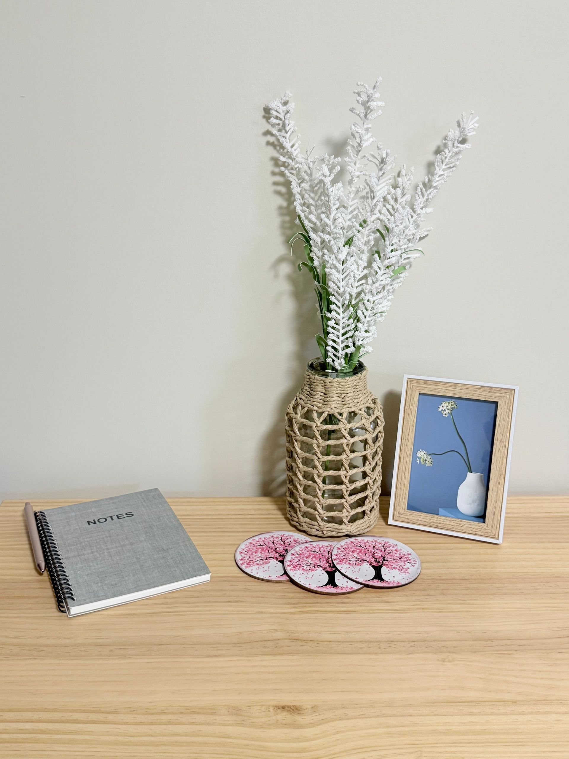 Woven vase with white flowers, framed photo, spiral notebook, and pink coasters on a wooden table