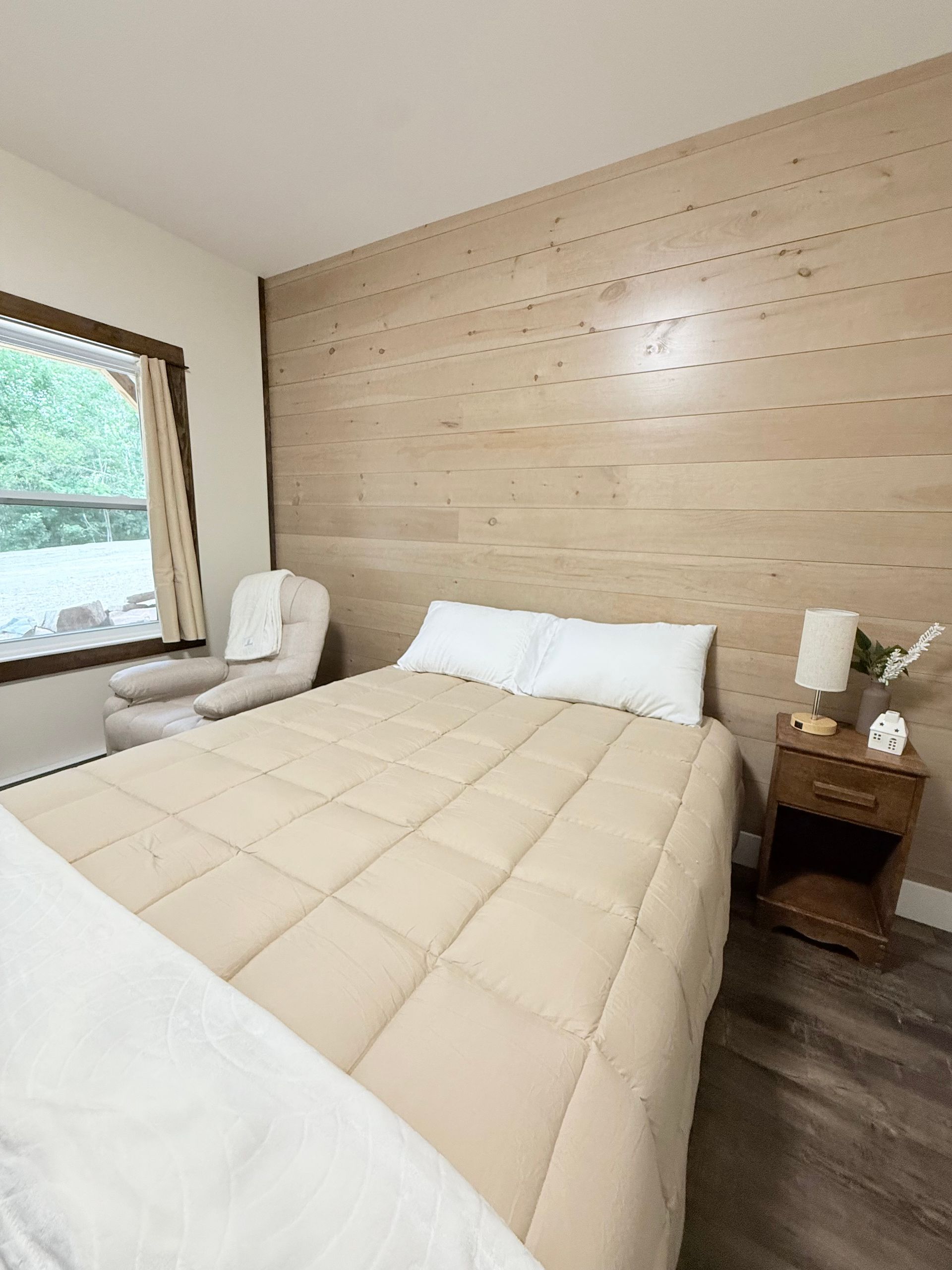 Neat bedroom with beige bed, wooden accent wall, white chair, and window view.