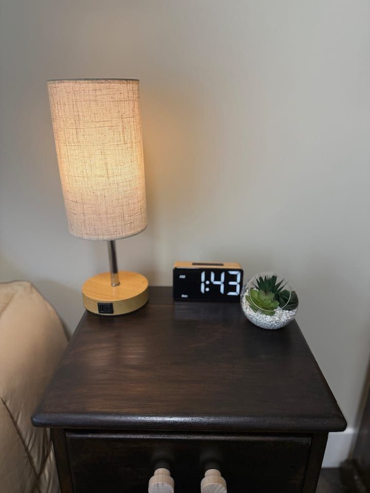 Small dark wood nightstand with a lit lamp, digital clock, and succulent plant beside a bed.