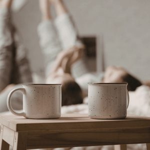 Two speckled mugs on a wooden table with a blurred person stretching in the background.