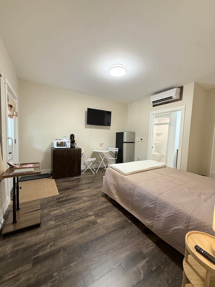Small bedroom with a bed, wall-mounted TV, mini fridge, desk, and air conditioner.