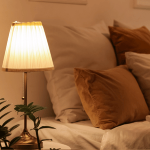 Warm bedroom scene with a lit bedside lamp beside beige pillows and a plant on the nightstand