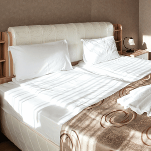 Neatly made bed with white linens, beige headboard, and a brown throw in a sunlit bedroom