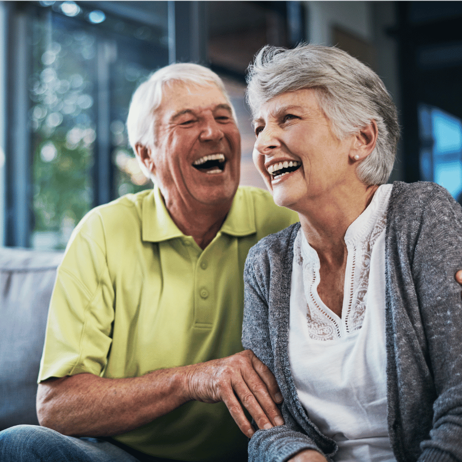 Smiling couple laughing together indoors, with the woman in a gray sweater and the man in a green polo.