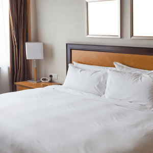 Bright hotel room with a neatly made white bed, wooden headboard, nightstand, lamp, and curtains.