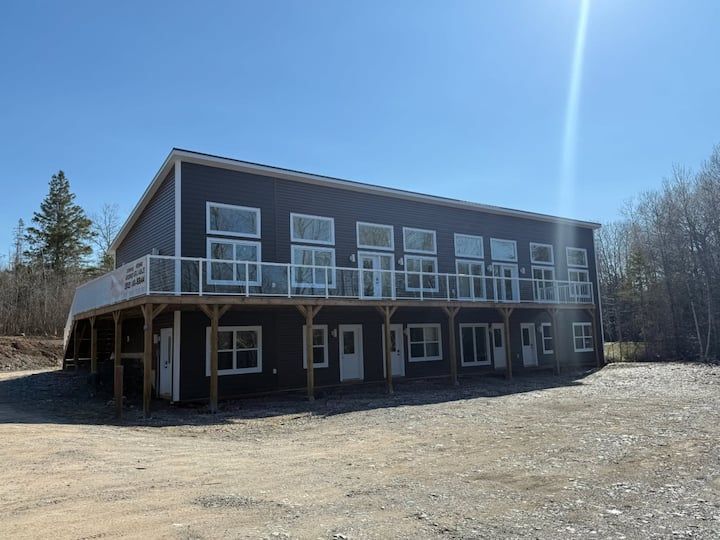 Two-story dark building with white-trimmed windows and a wraparound deck on a gravel lot under a clear blue sky