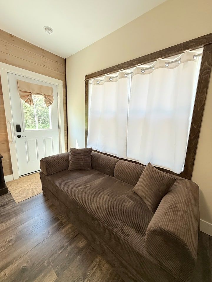 Small brown sofa in a bright entry room with white curtains and a front door.