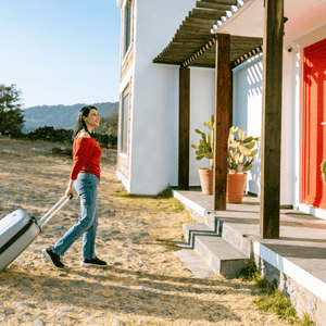 Woman pulling a suitcase toward a white house with a red door in a sunny yard