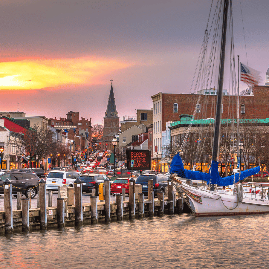 Harbor at sunset with docked sailboats, city street, and church spire in the background