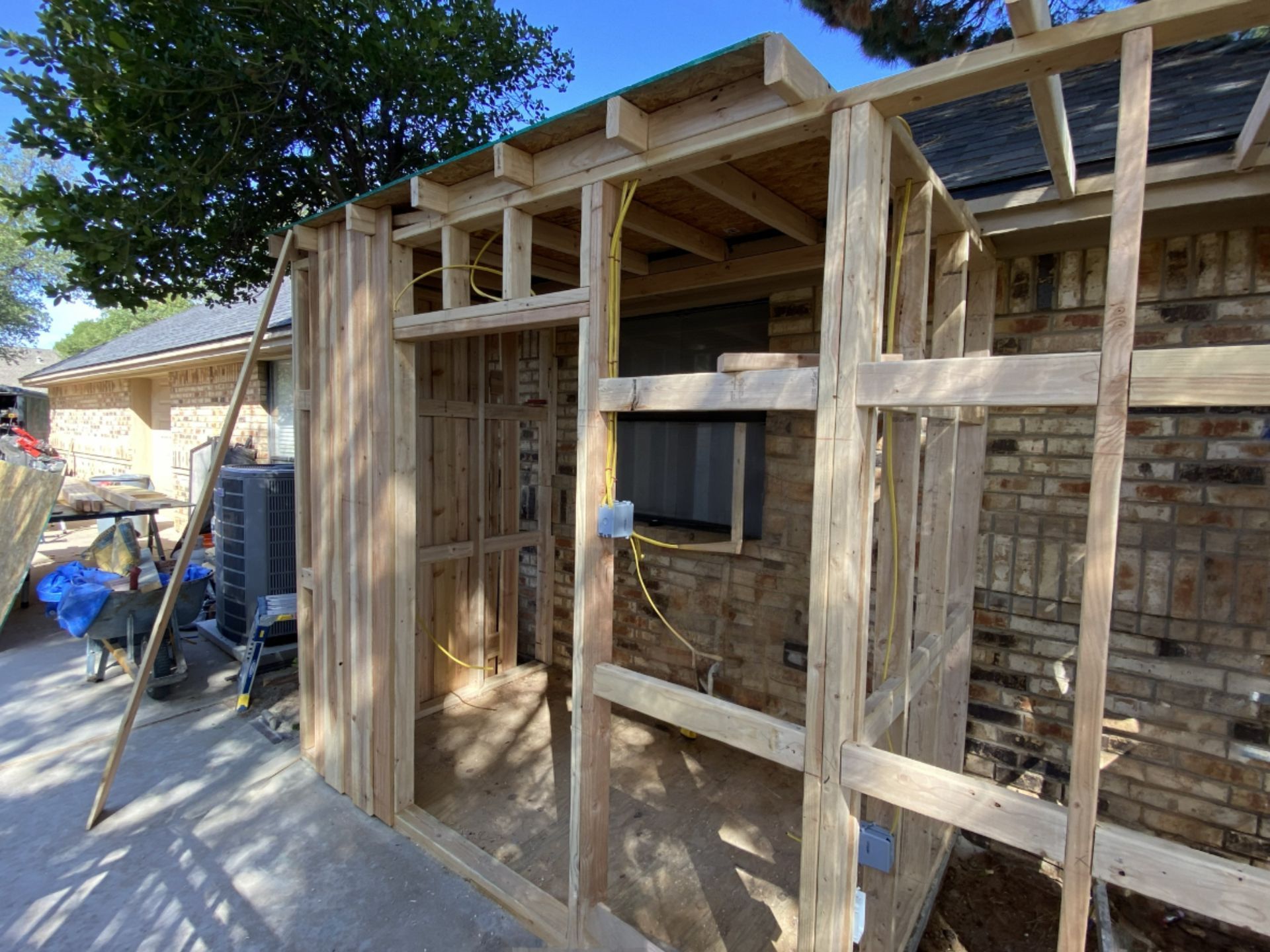 Wooden frame structure being built against a brick wall, with exposed wiring and partially completed roof.