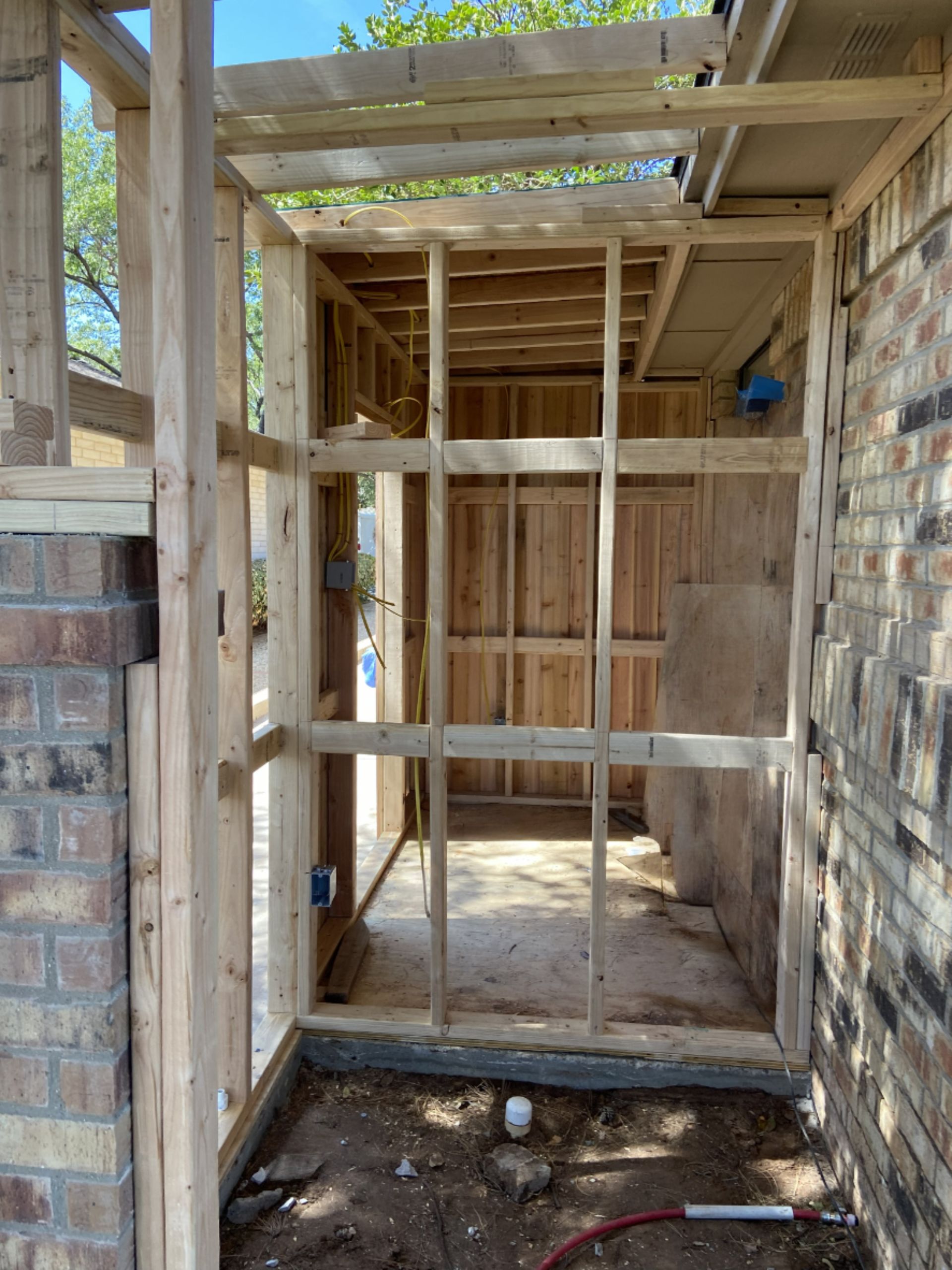 Wooden frame construction against a brick wall, possibly for an outdoor structure, with exposed studs and wiring.