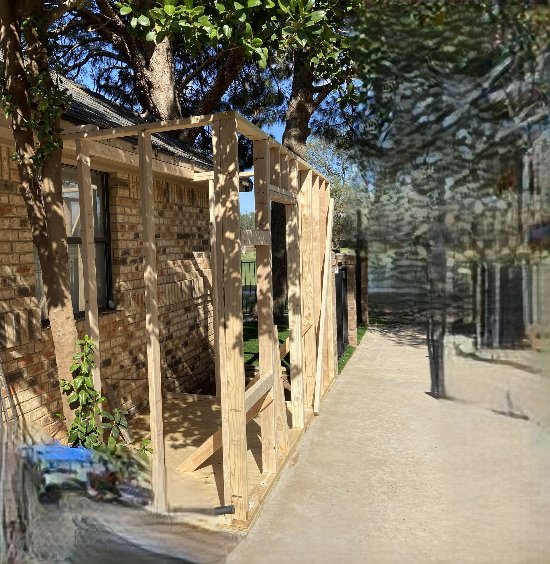 Wooden frame construction beside a brick wall, next to a concrete path and greenery under a sunny sky.