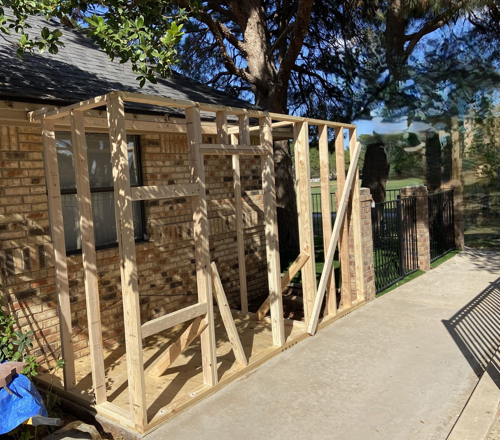 Wooden frame structure being built against a brick wall, on a concrete surface, near a tree.