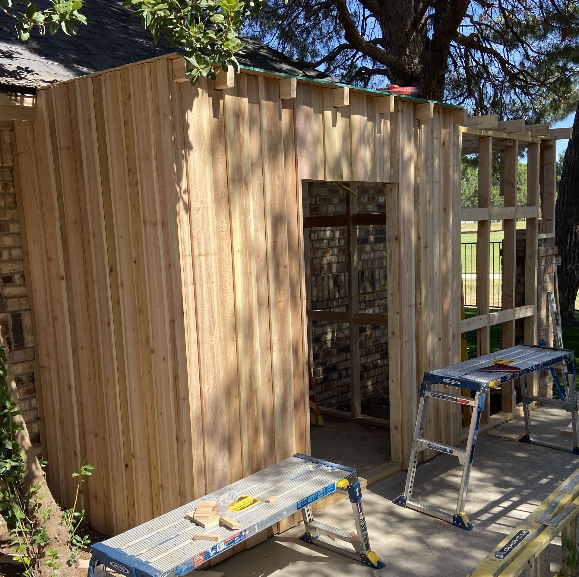 Wooden structure under construction outdoors with door frame, tools on ladders, and a brick wall.