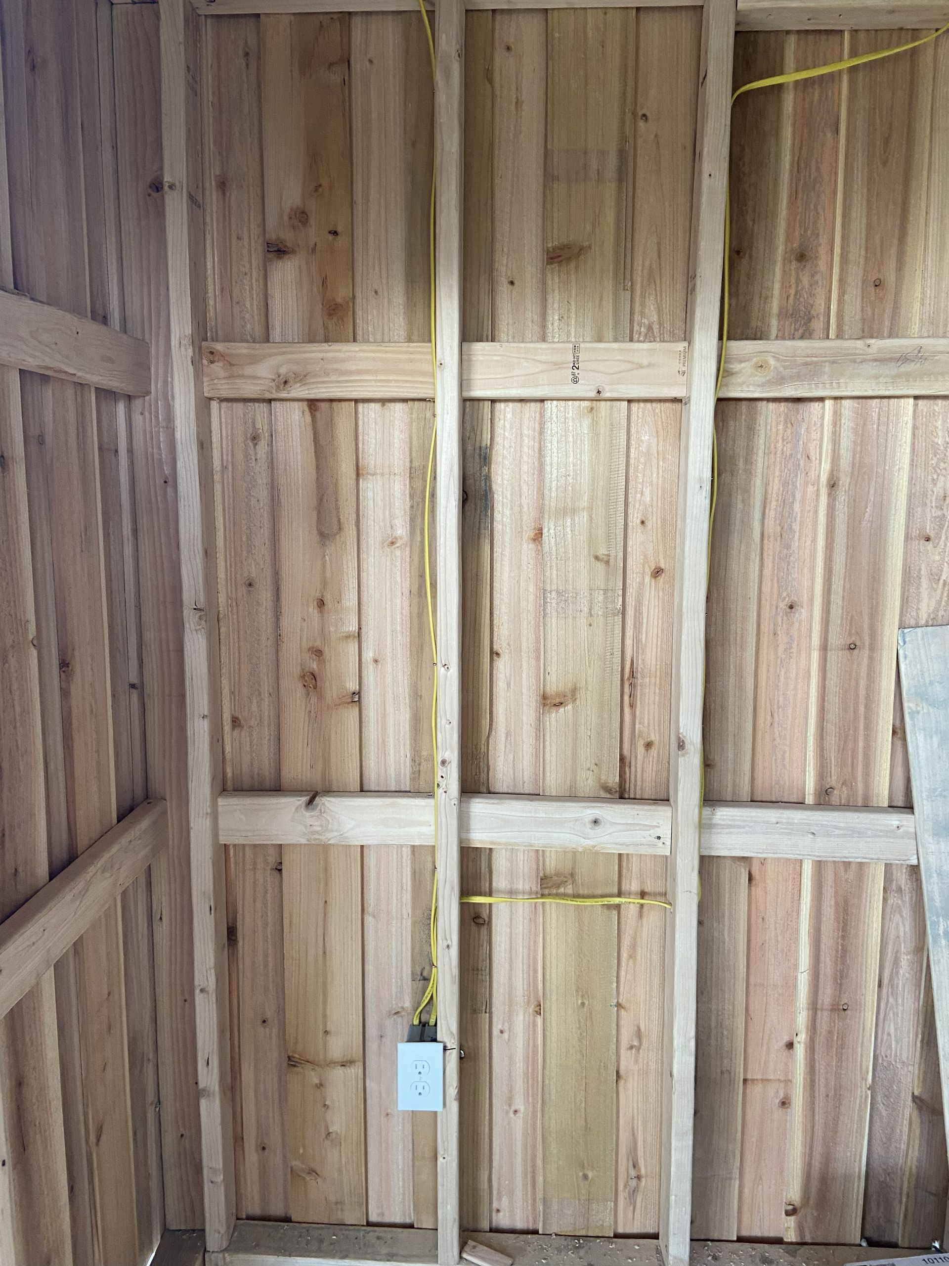 Interior wood-paneled wall with electrical outlet installed between wooden studs; yellow wire visible.