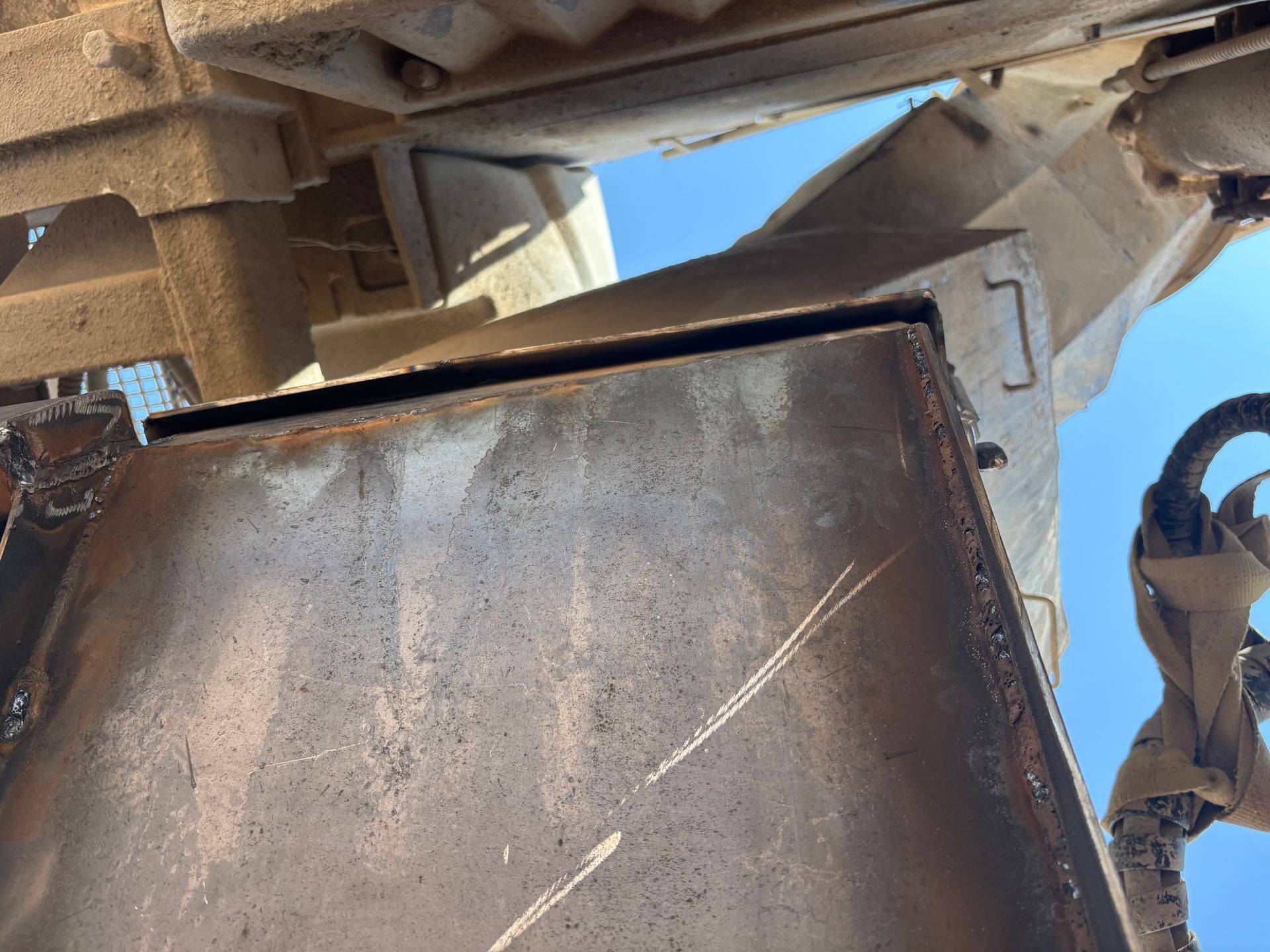 Close-up of a rusty metal box with scratches, connected to machinery against a blue sky.