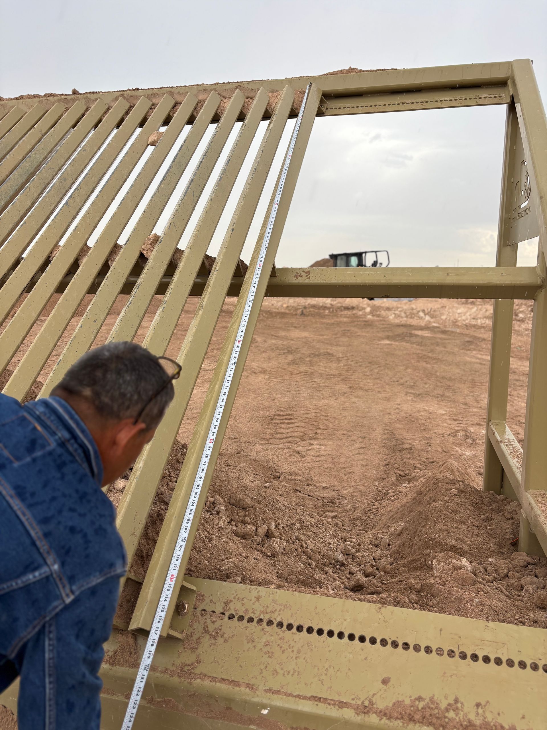 A person measures a metal frame structure with a measuring tape outdoors. A tractor is visible in the background.