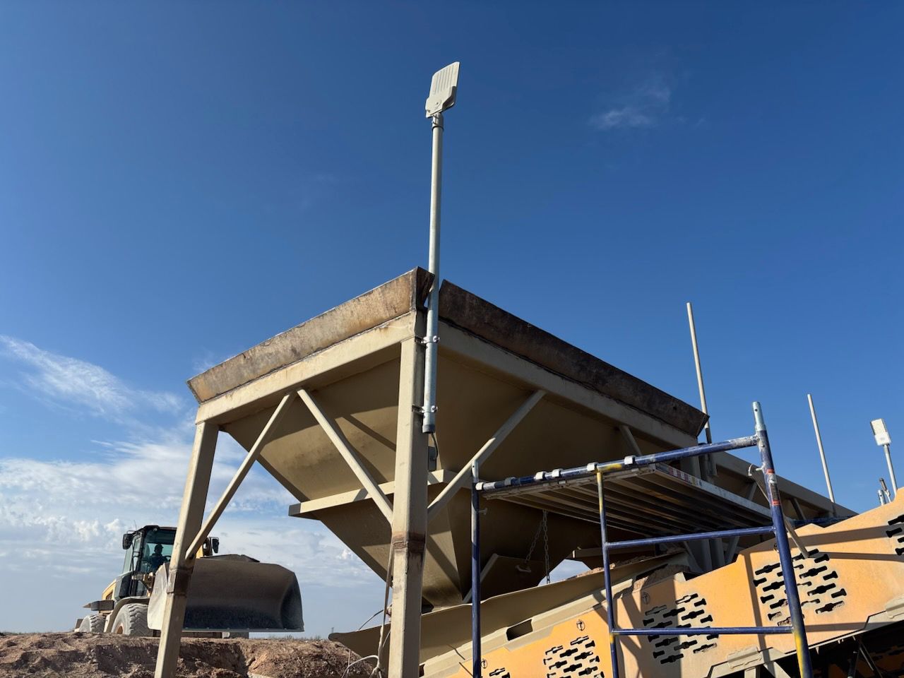 Construction site with industrial machinery under a clear blue sky. A tall light post is on top of a metal structure.