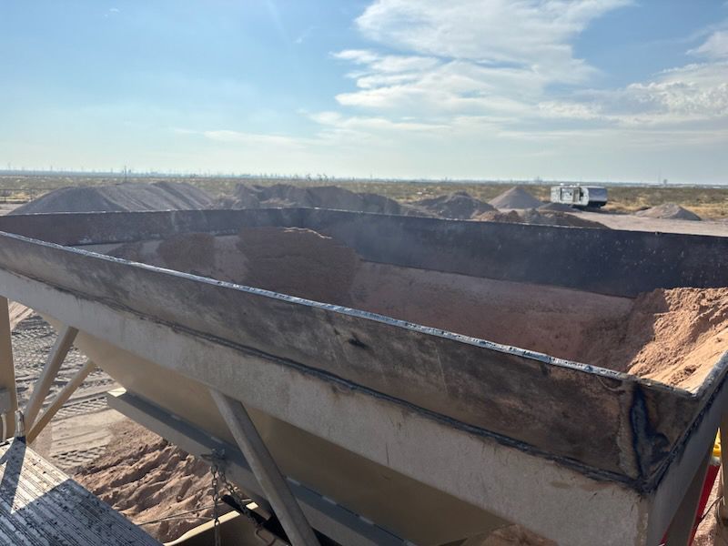 Large industrial bin filled with tan material; piles of material in the background under a blue sky.
