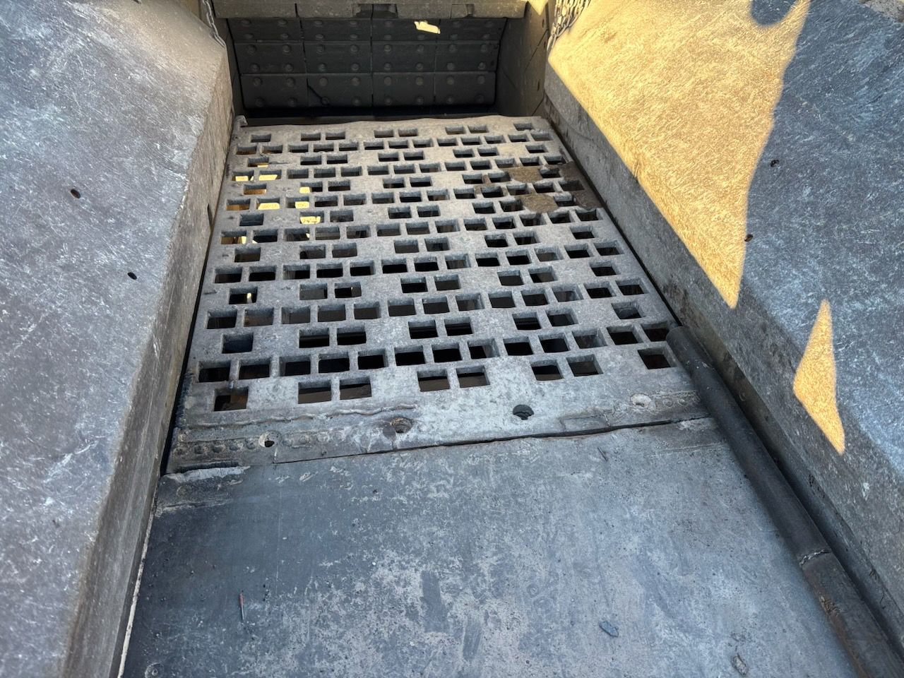 Interior view of a concrete pit with a square-holed metal grate. Harsh sunlight casts shadows.