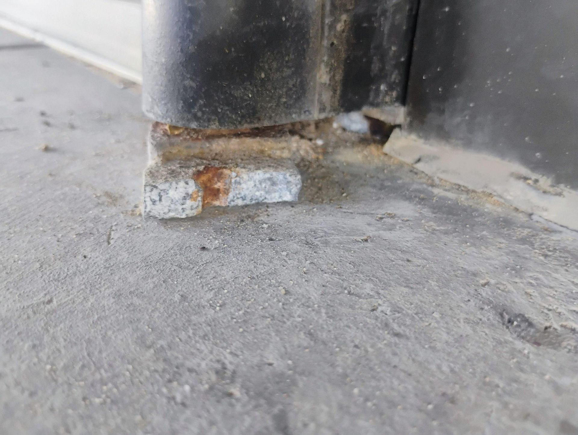 Close-up of a damaged metal door hinge on a concrete surface. Rust and debris are visible.