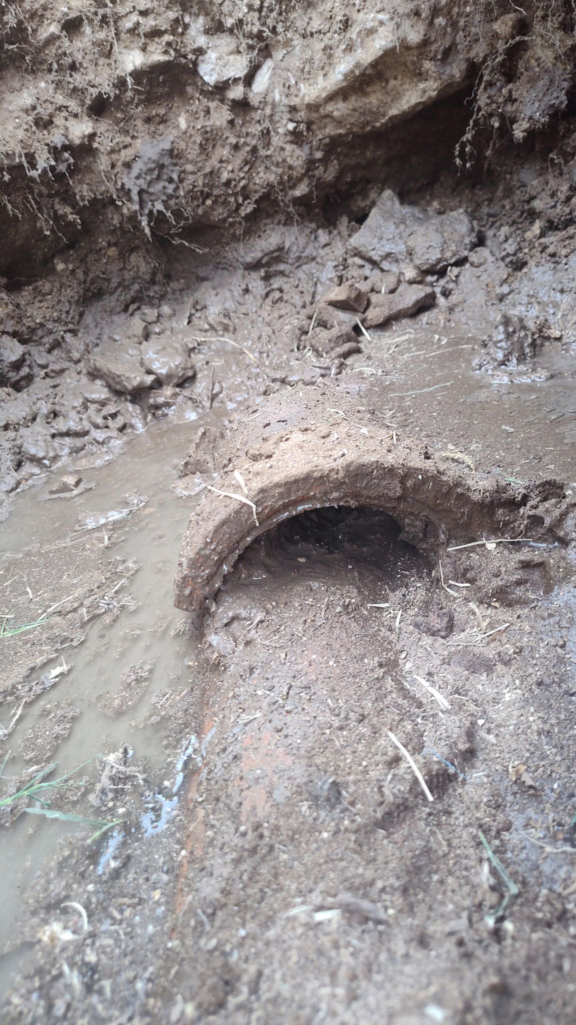 A dark, muddy trench with a section of round, dark pipe visible.