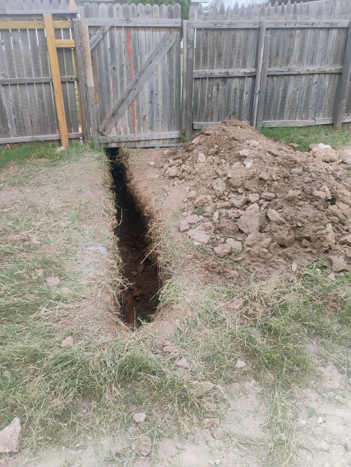 A trench dug in a backyard, with a pile of dirt beside it and a wooden fence in the background.