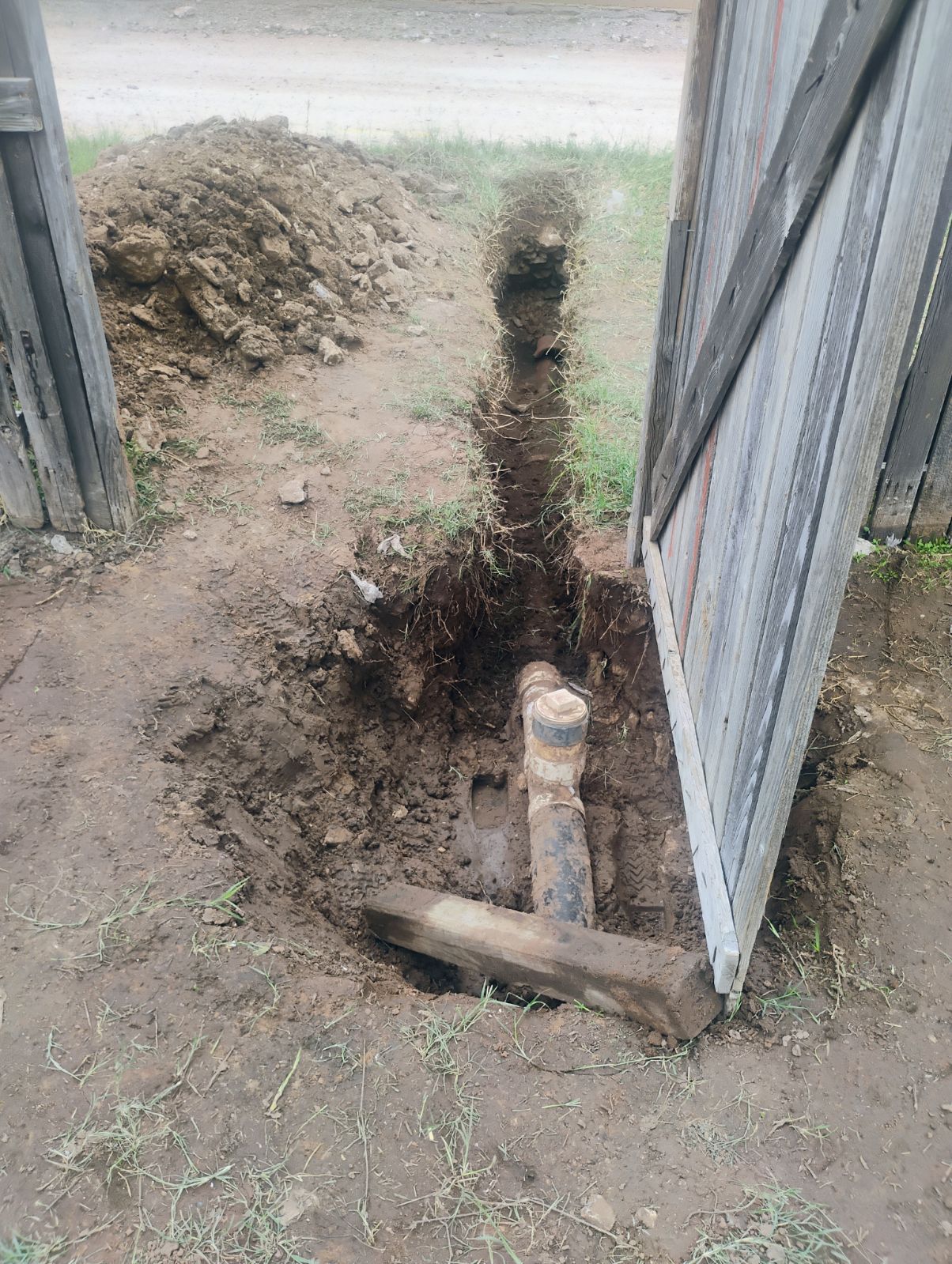 Trench dug near a fence with exposed pipes. Dirt pile in the background. Gate open.