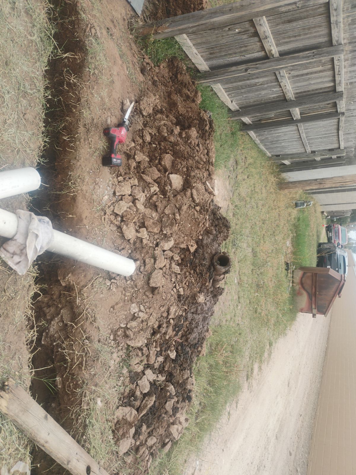 Trench in yard with white pipes, dirt, and tools, near a wooden structure.