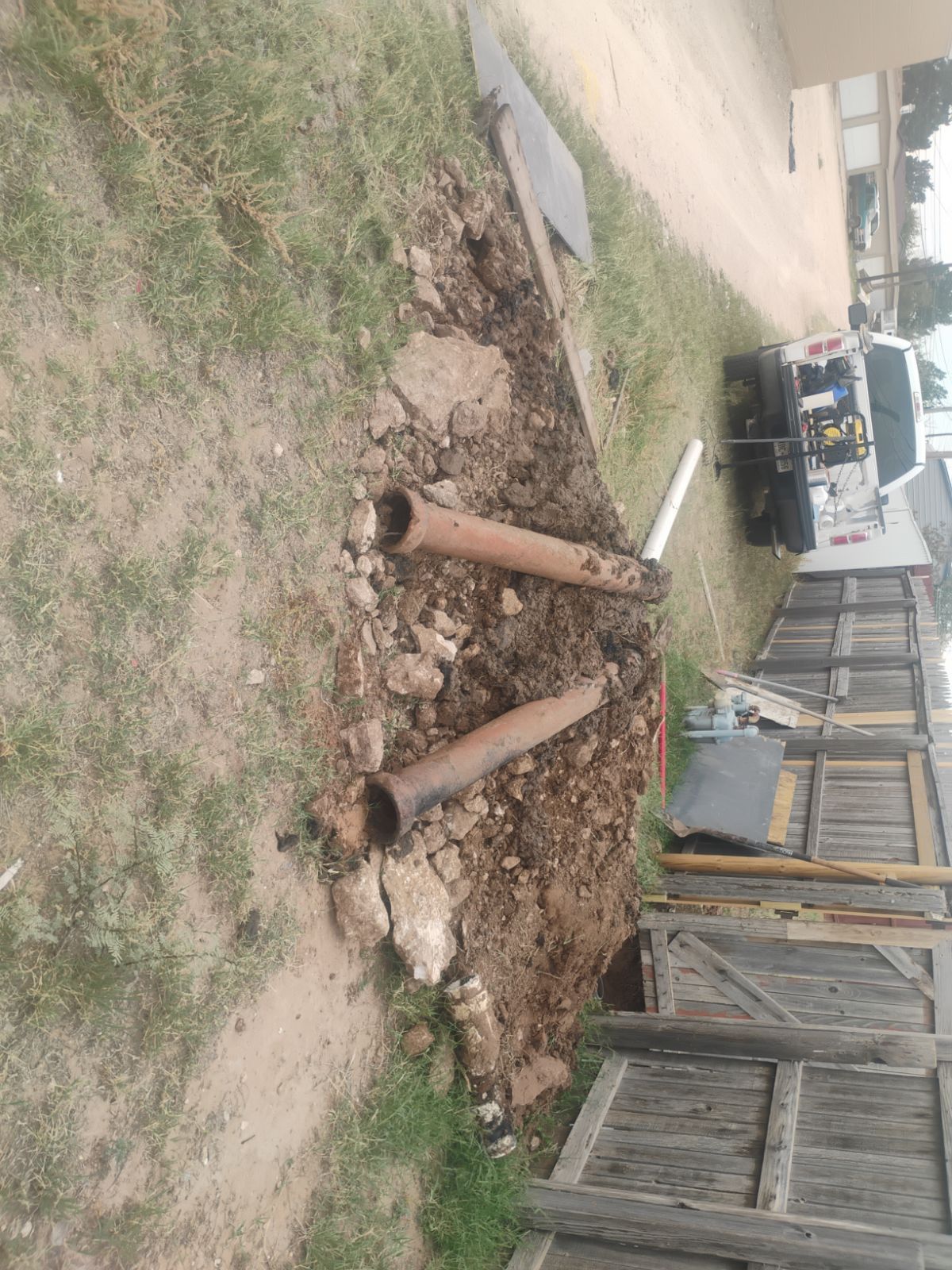 Excavation site with exposed pipes in a yard. A work truck is visible in the background.