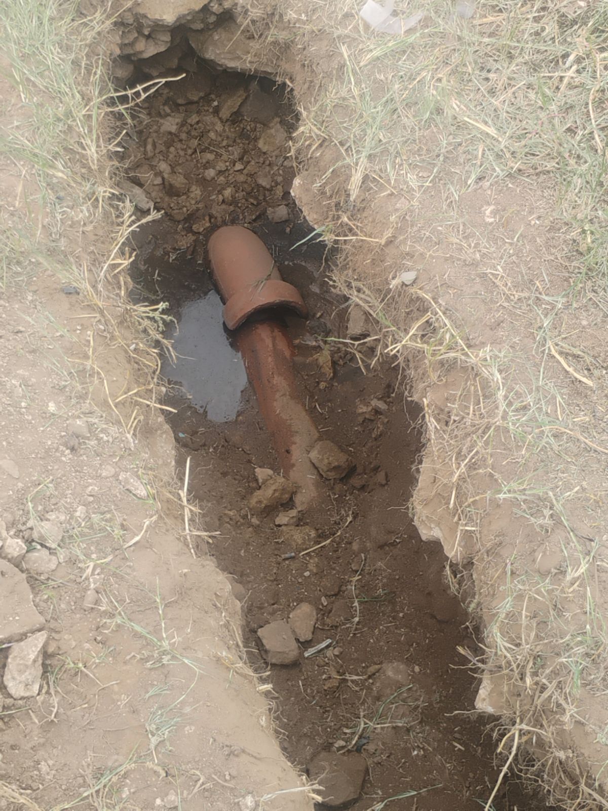 A rusted pipe with a leak exposed in an excavated trench, surrounded by dry earth.