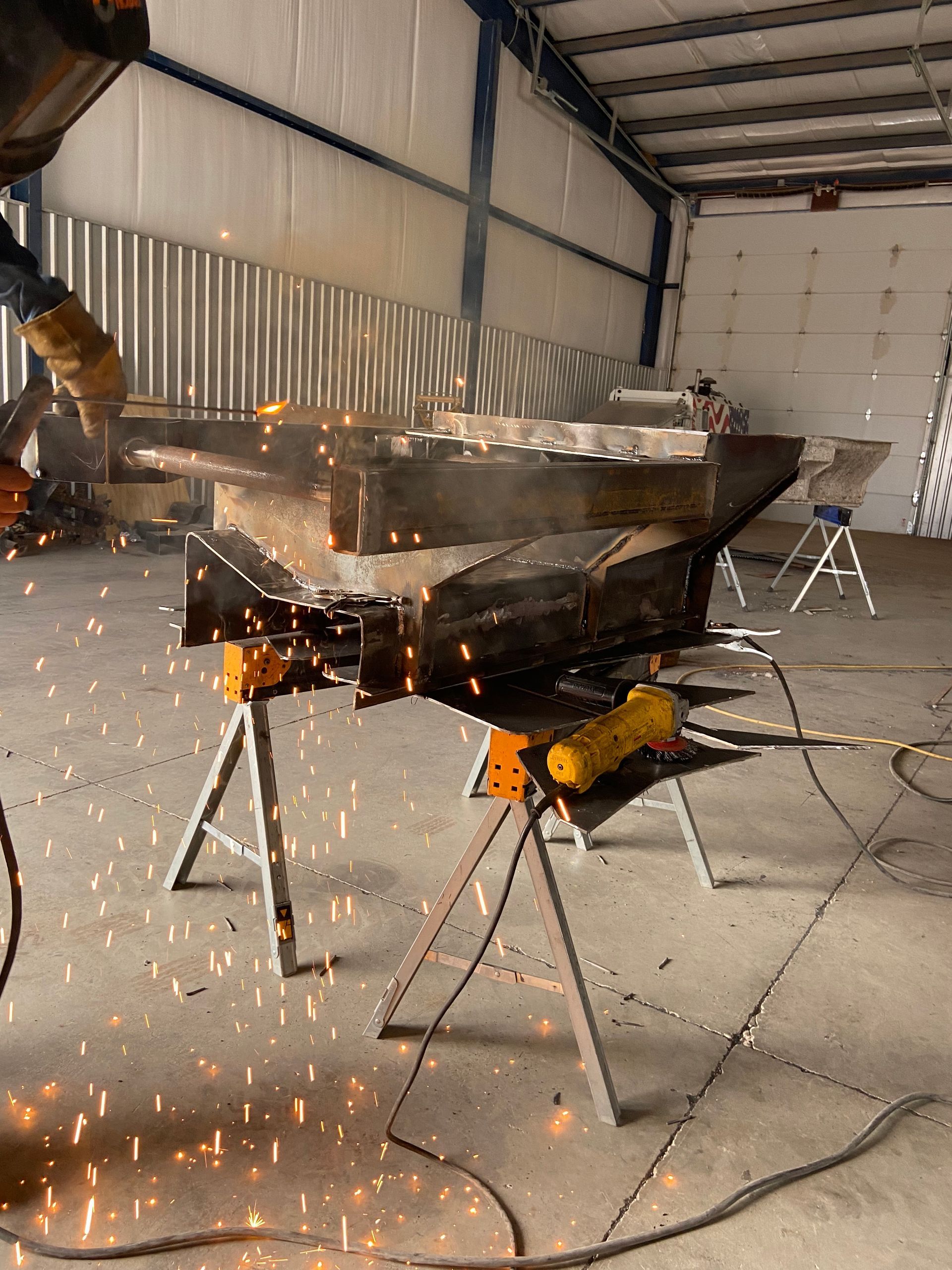 Sparks fly as someone welds metal structure in a workshop. Sawhorses support the work.