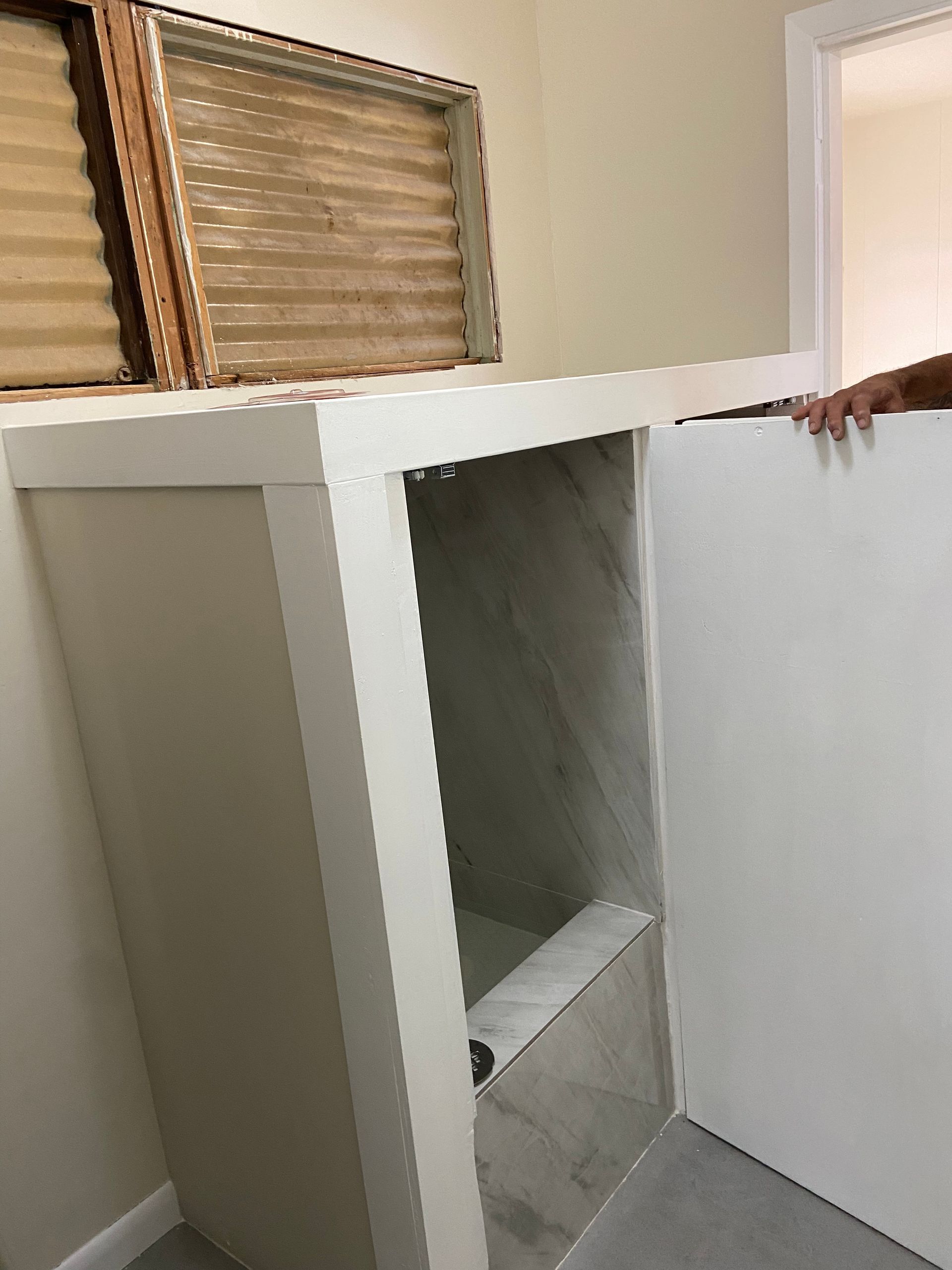 White cabinet with open door; person holding the door. Beige walls, window, and gray flooring.