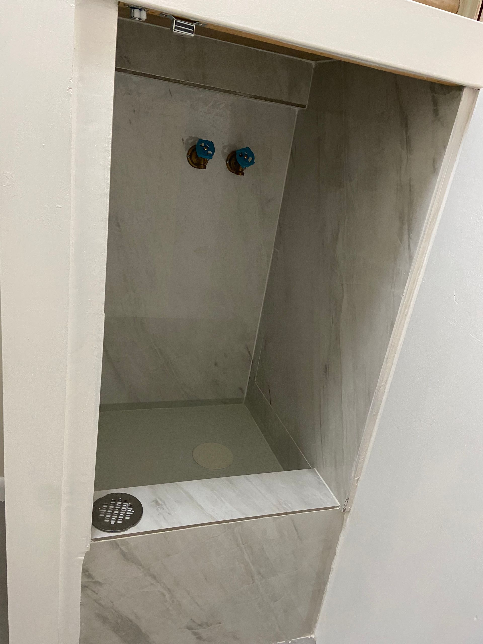 Recessed, tiled shower with blue water taps and a drain in the bottom. White frame.