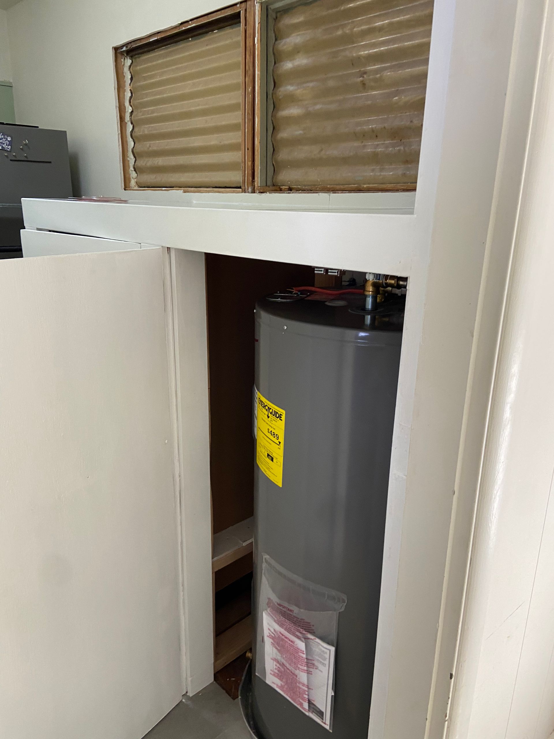 A water heater inside a closet; a white door is open. Above is a window with blinds.