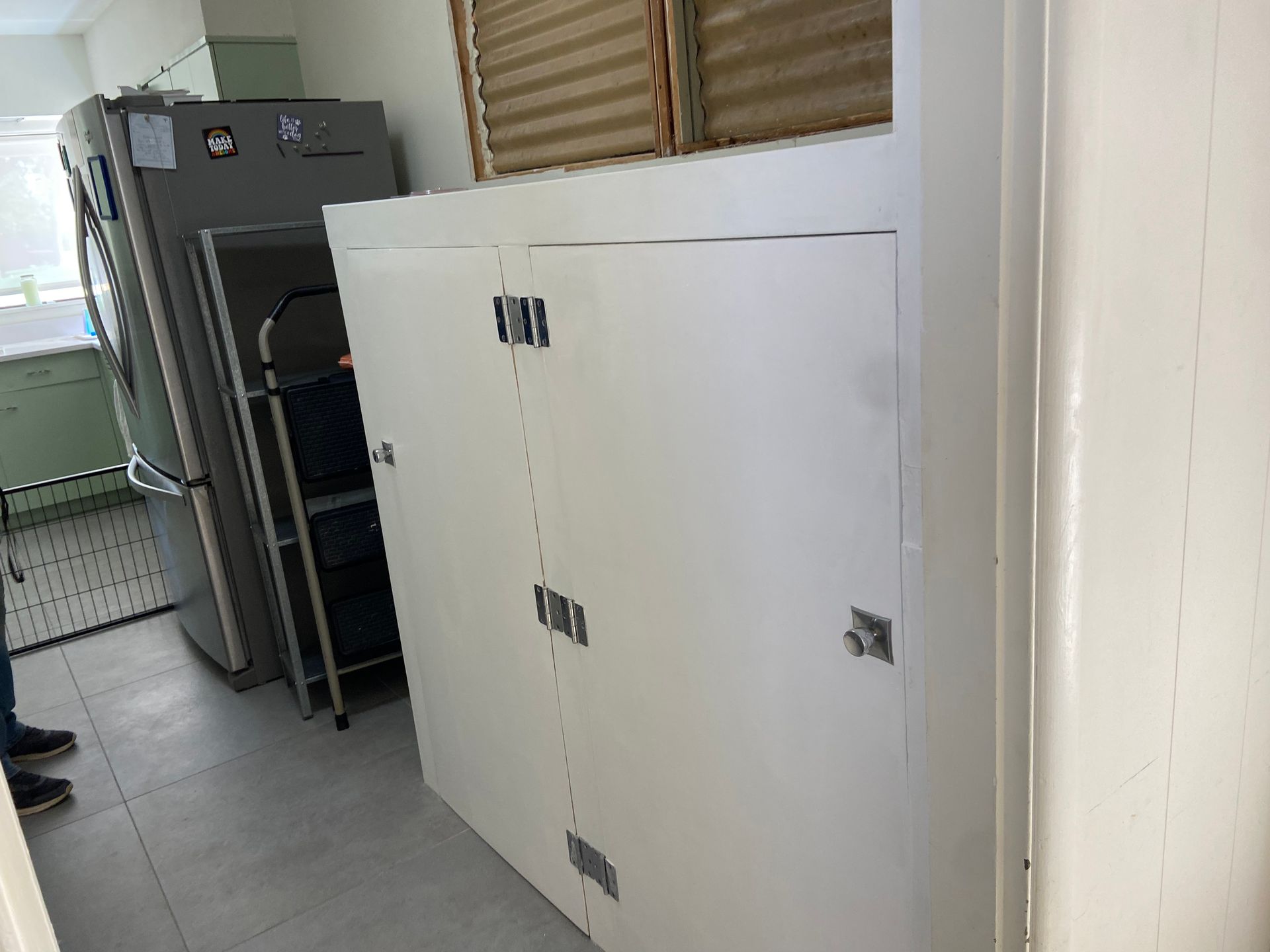 A white cabinet with two doors and silver latches, next to a refrigerator and shelves in a kitchen.