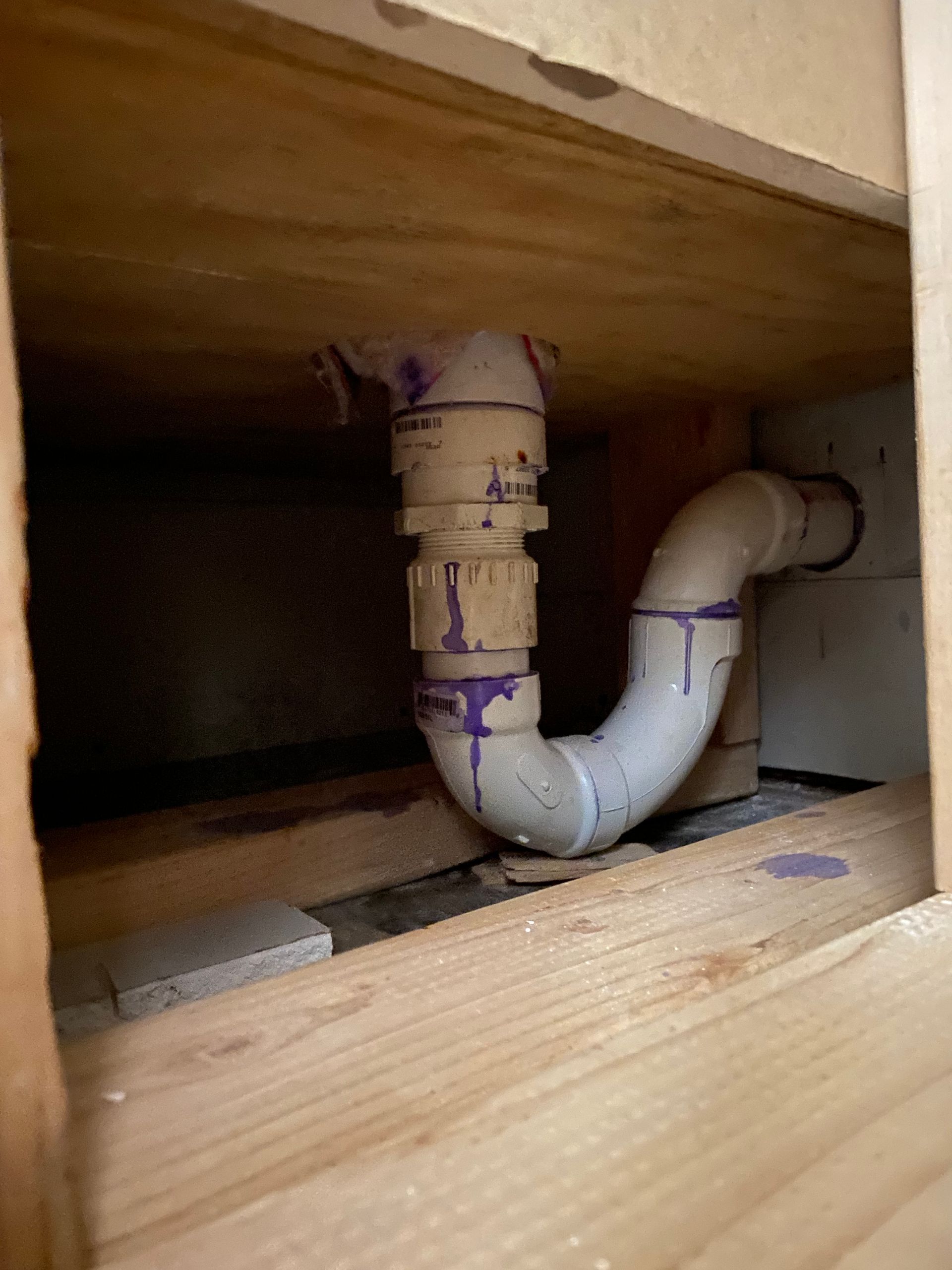 White PVC plumbing trap inside a wooden cabinet frame, purple glue visible.