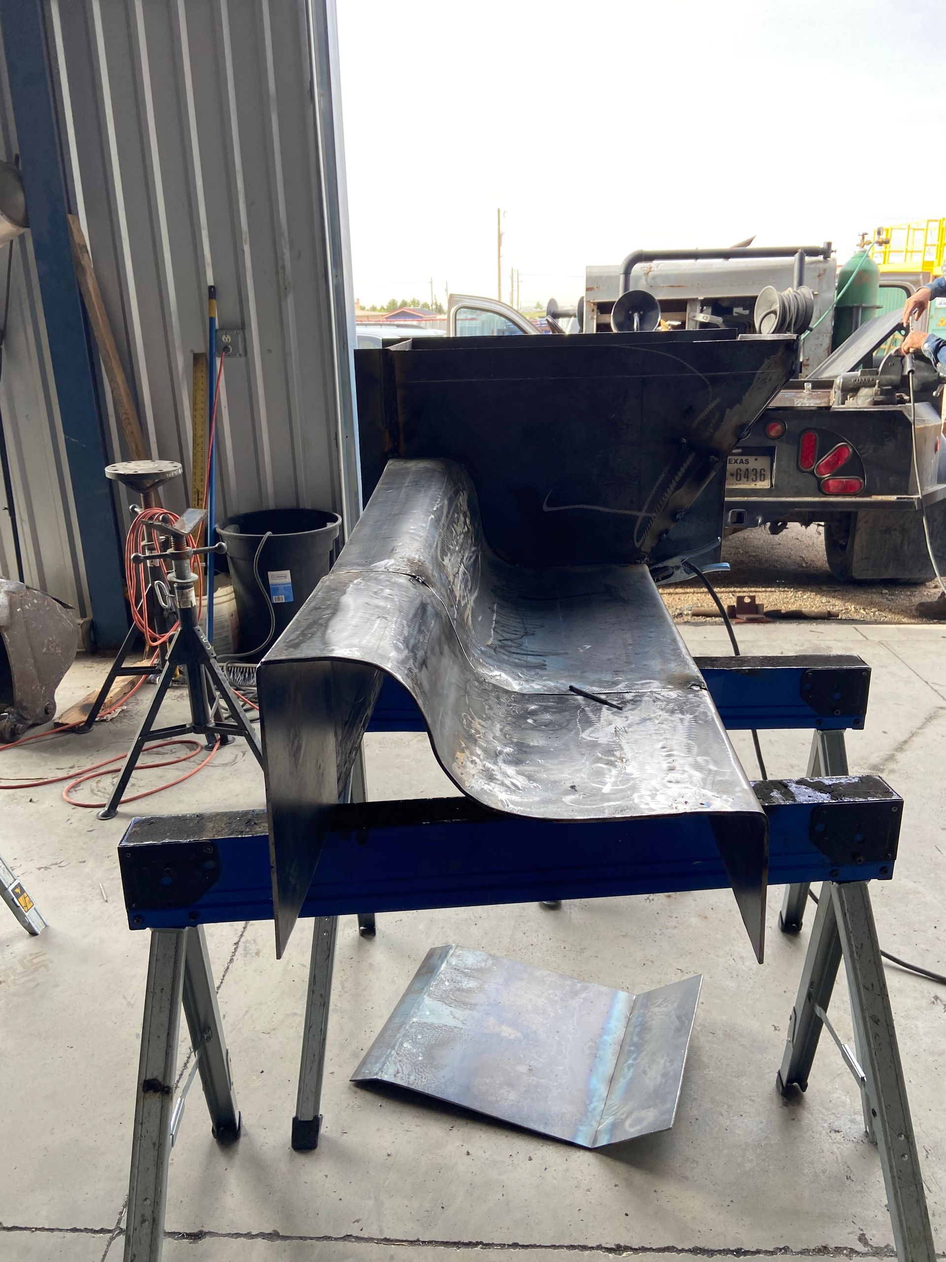 Metal work in progress on sawhorses outside a garage. A large, curved sheet is being worked on.