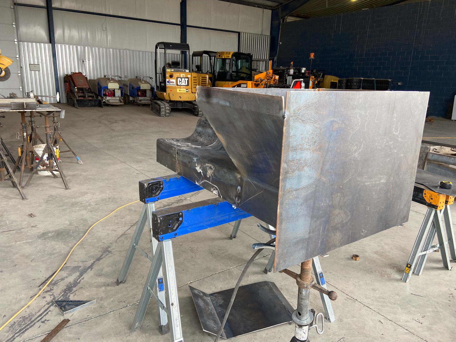 Welded metal structure on blue sawhorses in a workshop with heavy machinery in the background.