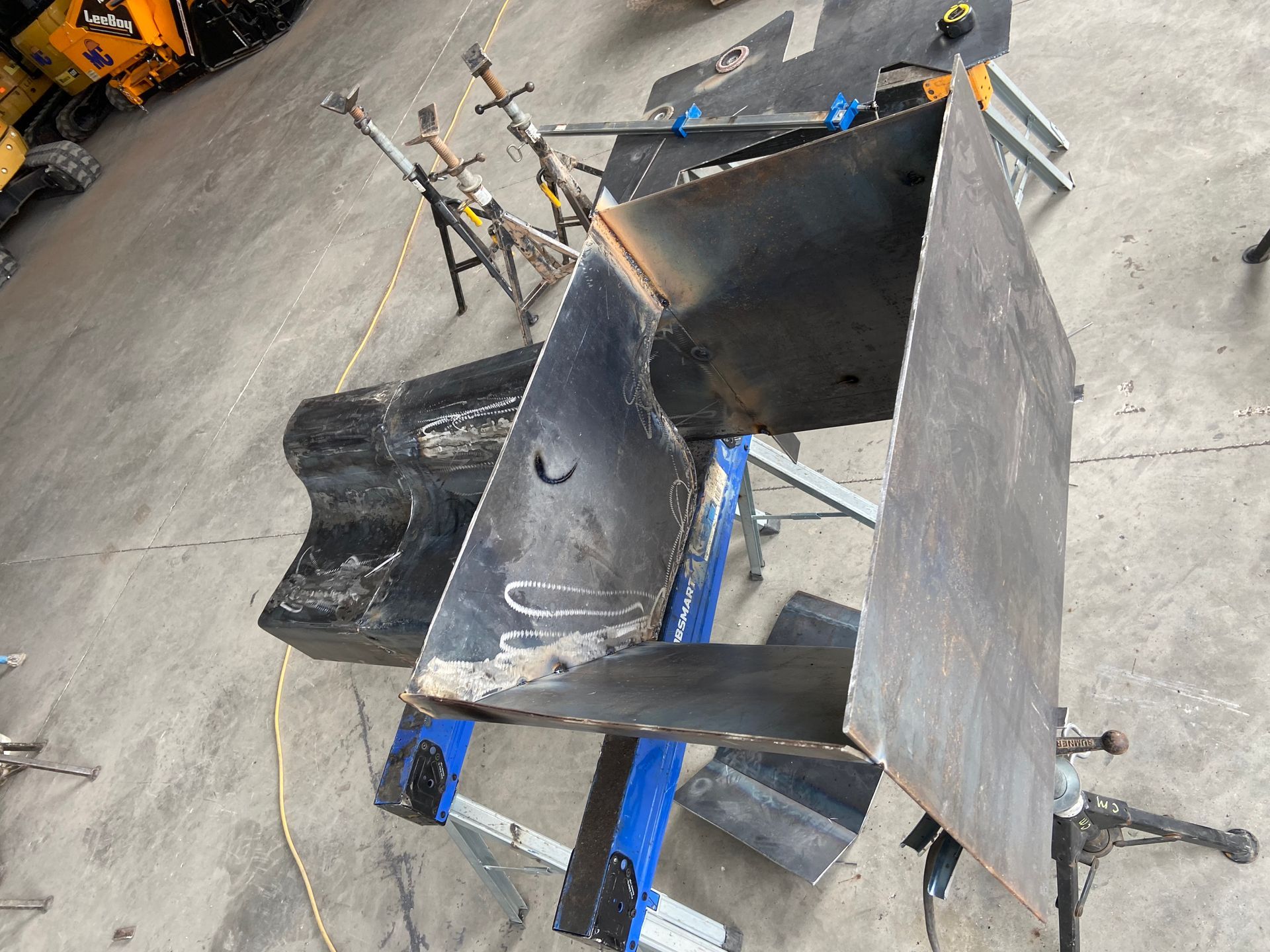 Metal sheets, welded, forming a structure, on a workbench. Blue clamps visible.