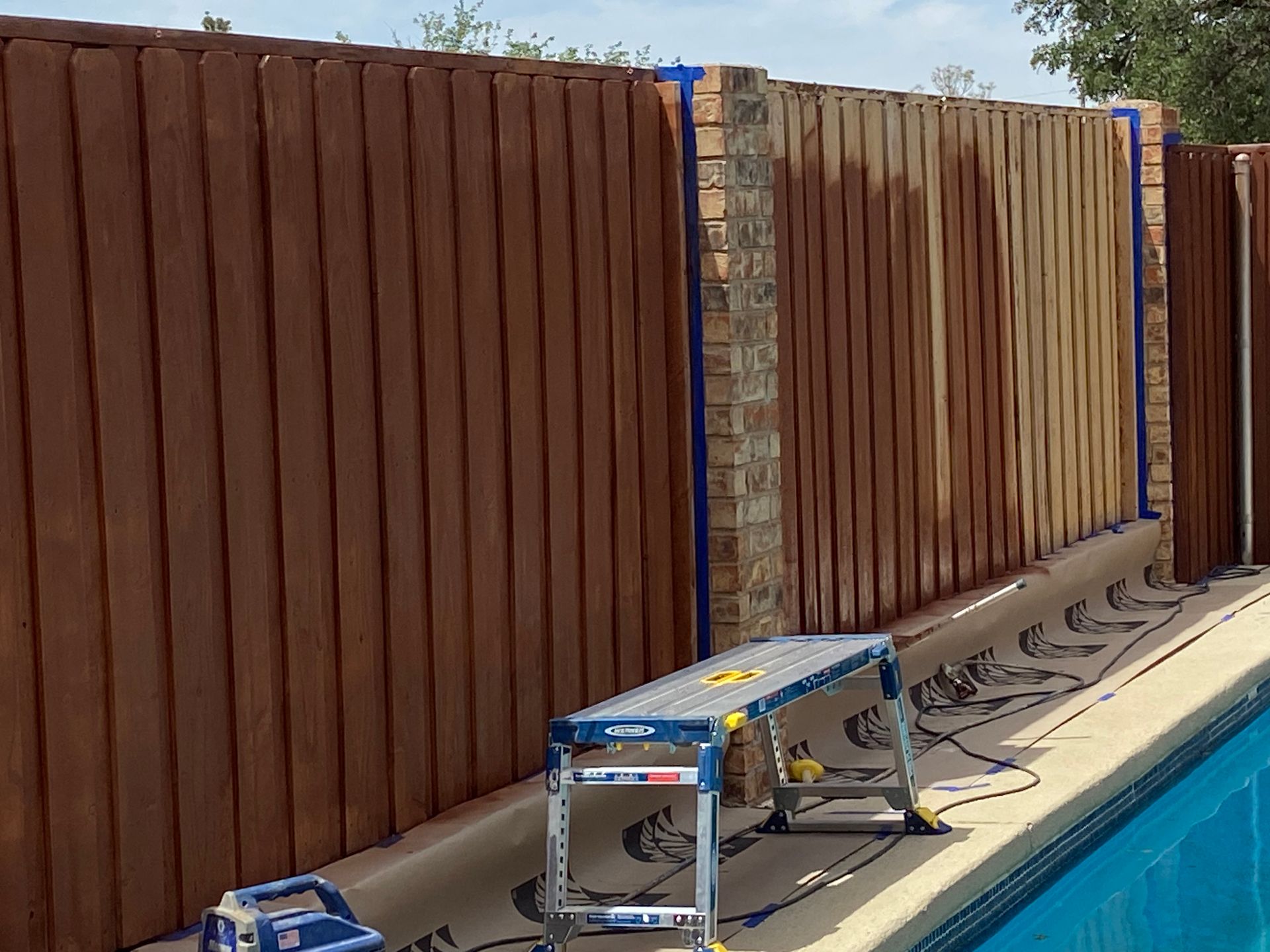 Brown wooden fence next to a pool being painted. A ladder is set up along the wall.